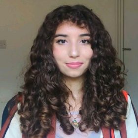 This is a coloured picture of Val Saddi. Val has shoulder-height golden brown curly hair. She is wearing a mesh top with a black vest underneath. She is also wearing a striped blazer, with orange, brown and white stripes alternating. Val is a white mediterranean woman with olive skin. She has dark brown eyes, a straight nose, plump pink lips. She is smiling in this picture.